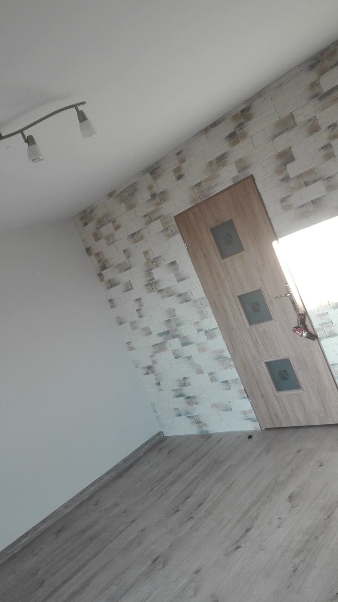 Wnętrze po remoncie: drewnopodobna podłoga, ściana z dekoracyjną okładziną imitującą cegłę, zamontowane drzwi wewnętrzne z trzema szybkami, widoczna wkrętarka na podłodze.