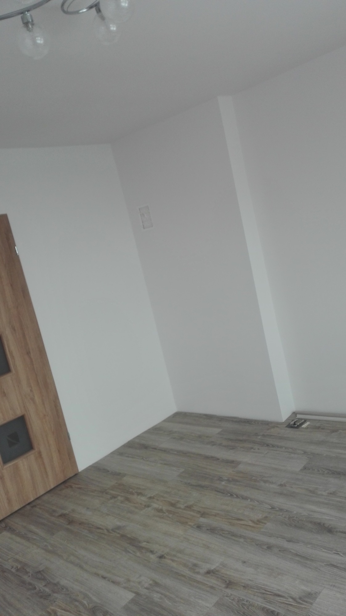 Nowo wykończone wnętrze z drewnopodobnymi panelami podłogowymi, białe ściany i częściowo widoczne drzwi z drewnianą okleiną i czarnymi intarsjami, widoczna kratka wentylacyjna w podłodze przy ścianie.