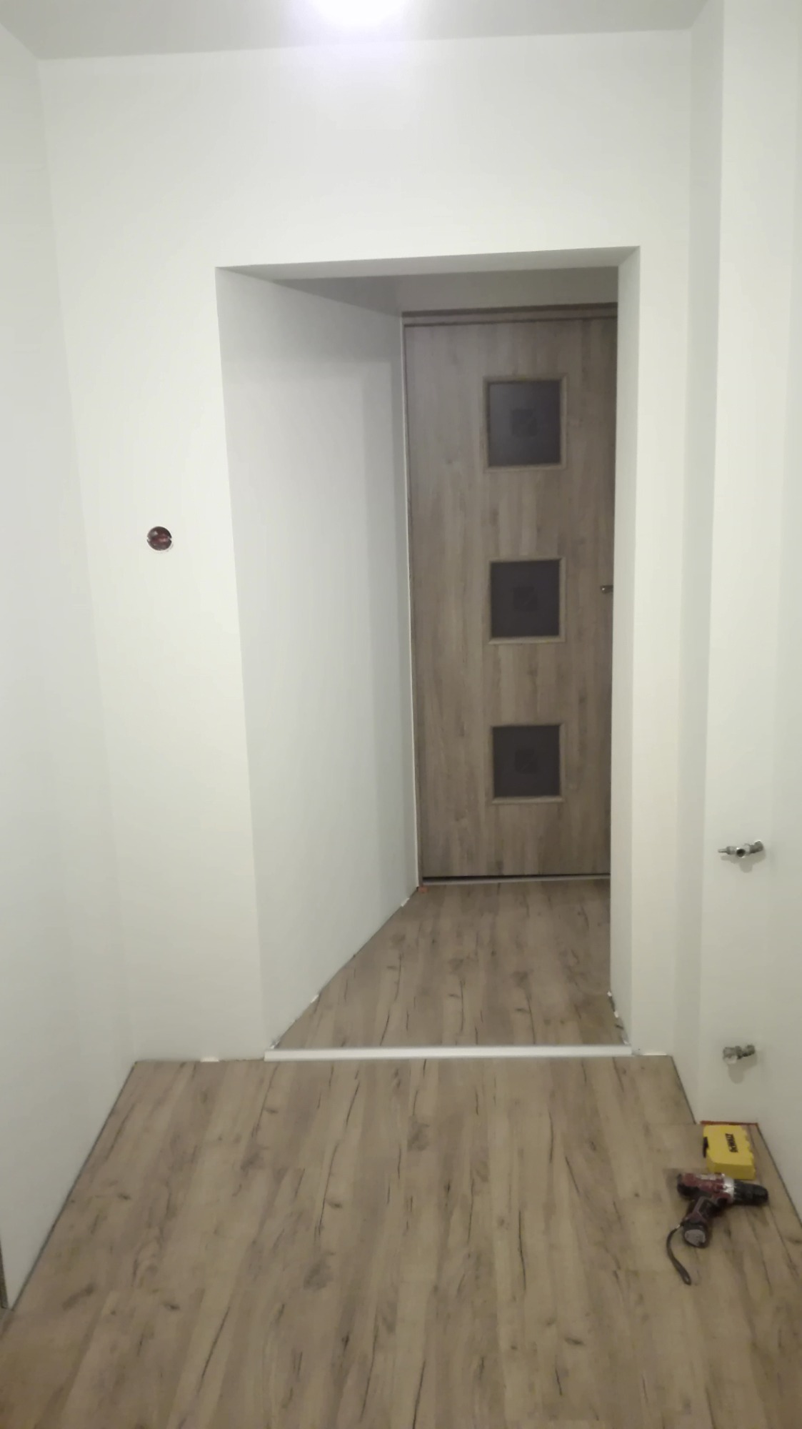 Wnętrze po remoncie: świeżo położone panele podłogowe w odcieniach brązu, białe ściany z widocznym otworem drzwiowym i zamontowanymi drzwiami wewnętrznymi z trzema kwadratowymi przeszkleniami,...