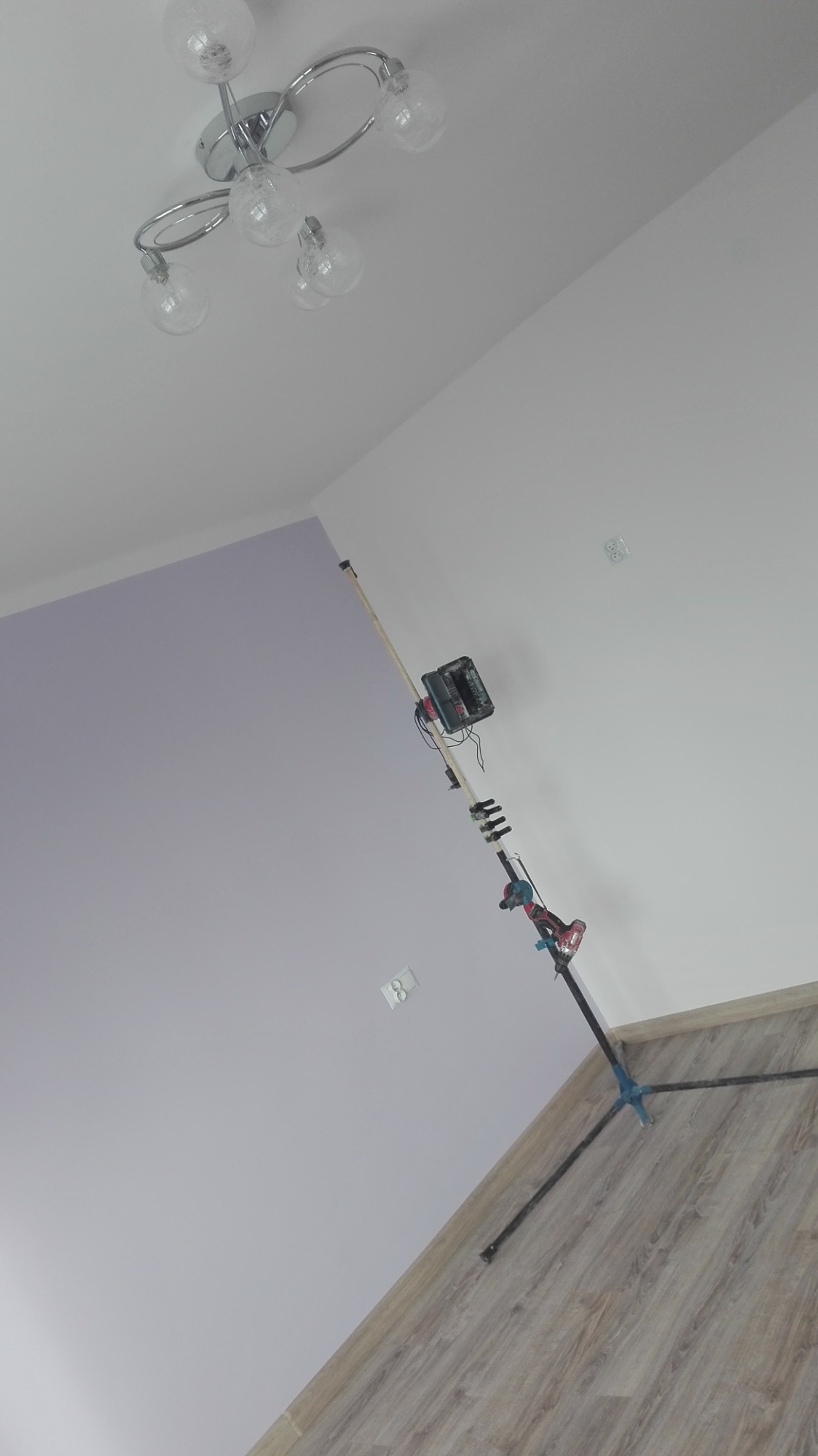Wnętrze w trakcie prac wykończeniowych: widoczny statyw z narzędziem do szlifowania ścian pod kątem, ściana pomalowana częściowo na fioletowo, częściowo na biało, drewnopodobna podłoga oraz lampa...