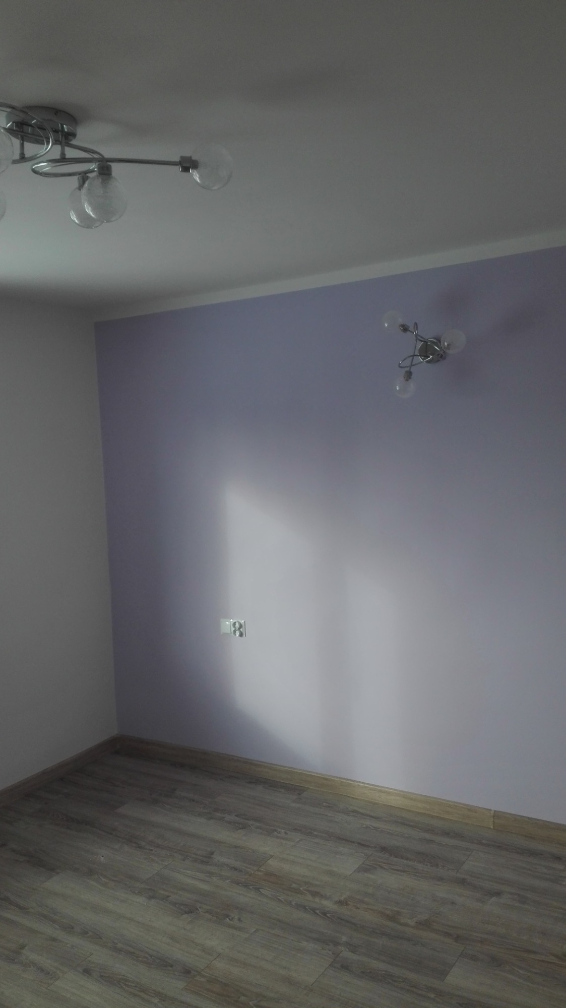 Pokój po remoncie: świeżo pomalowane ściany w odcieniach fioletu, ułożone panele podłogowe imitujące drewno, widoczne gniazdko elektryczne i dwa różne modele lamp sufitowych.