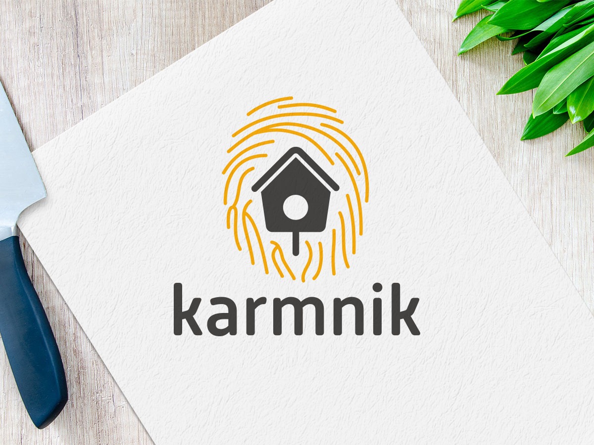 Projekt logo 'karmnik' z motywem ptasiej budki w odcisku palca, leżący na papierze obok noża i zielonych liści.