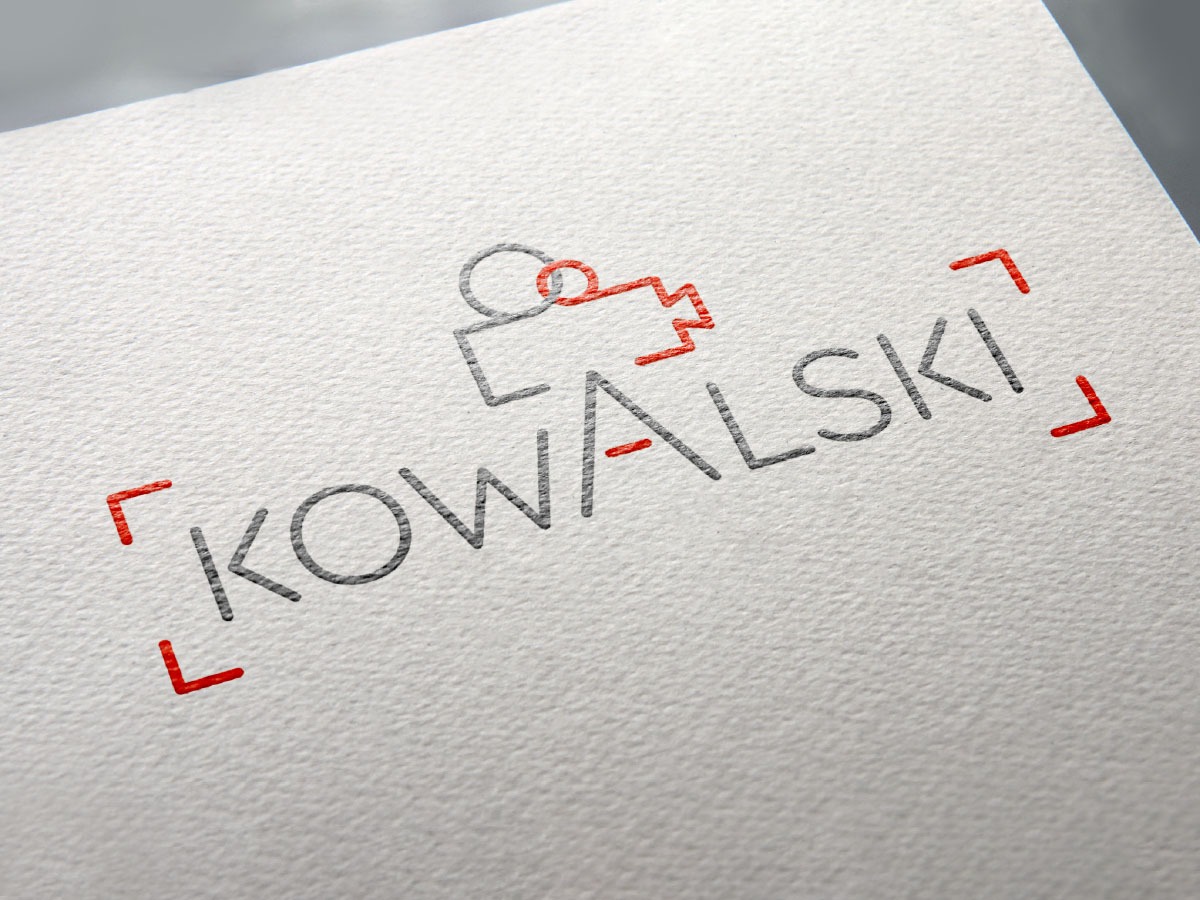Minimalistyczny projekt logo z nazwą 'KOWALSKI' w szarym kolorze, ujętą w czerwone nawiasy, oraz graficznym symbolem w górnej części logo, umieszczony na teksturowanym, białym papierze.