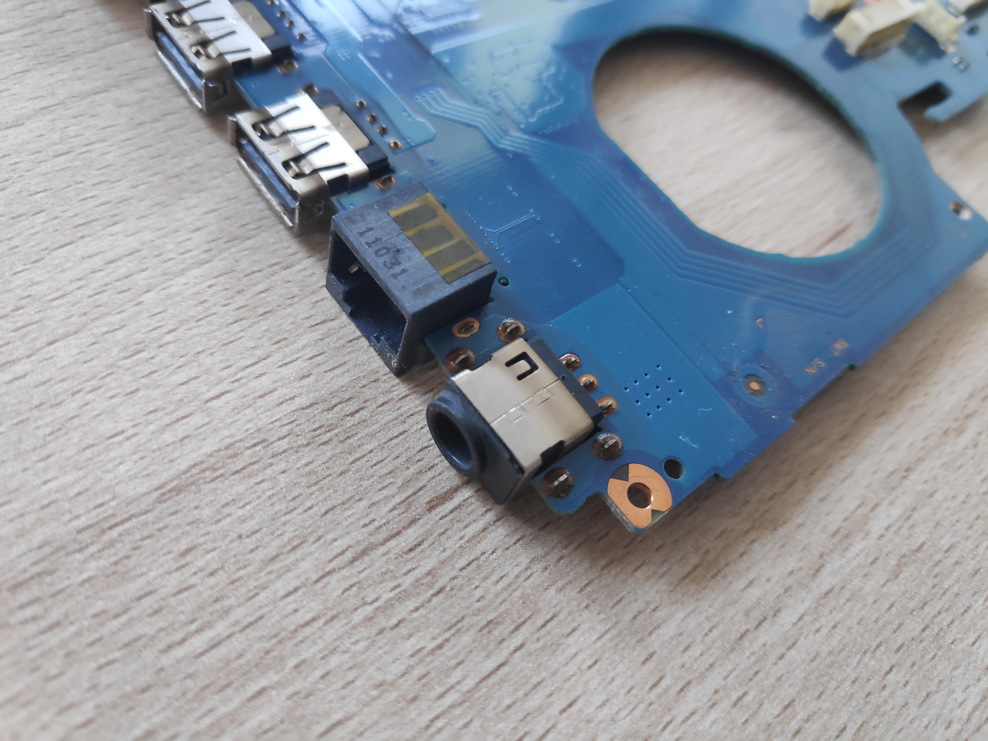 Fragment niebieskiej płyty głównej laptopa z widocznymi portami USB oraz gniazdem zasilania, leżący na jasnym drewnianym blacie.