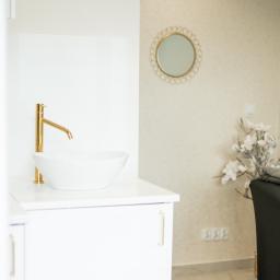 Avenue Estetic Klinika Kosmetologii - Jasne wnętrze gabinetu z umywalką z pozłacaną baterią i okrągłym lustrem w złotej ramie, obok leżanka kosmetyczna z ręcznikiem i dekoracją kwiatową.