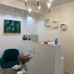 Recepcja kliniki z białym biurkiem, laptopem, kwiatami w wazonie, zielonym krzesłem, obrazem z pawimi piórami i designerskim żyrandolem.