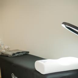 Avenue Estetic Klinika Kosmetologii - Przygotowane stanowisko zabiegowe z czarnym leżanką, białą poduszką ortopedyczną, złożonym szarym ręcznikiem i lampą pierścieniową w tle jasnych ścian i dekoracyjnych kwiatów.