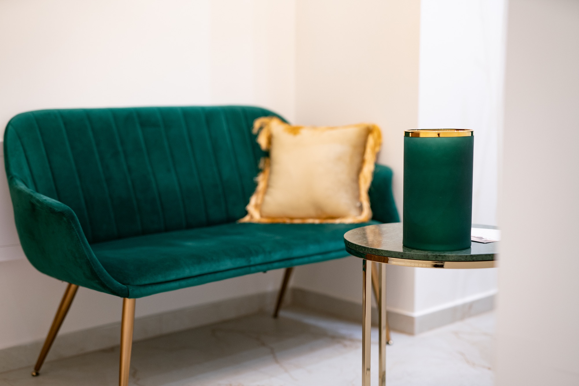 Szmaragdowa sofa z weluru z poduszką w kolorze złota, okrągły stolik z zielonym blatem i złotymi nogami, na którym stoi matowy, zielony wazon ze złotym obramowaniem, marmurowa podłoga.