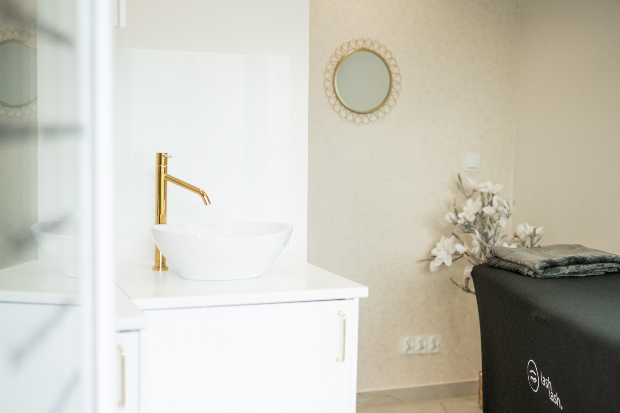 Jasne wnętrze gabinetu z umywalką z pozłacaną baterią i okrągłym lustrem w złotej ramie, obok leżanka kosmetyczna z ręcznikiem i dekoracją kwiatową.