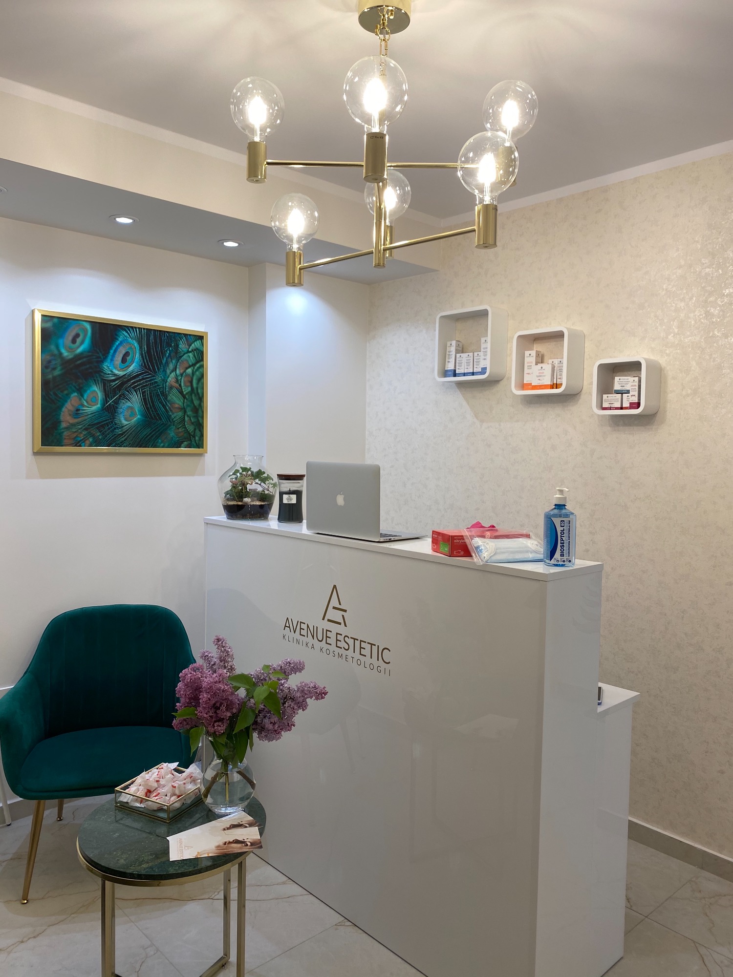 Recepcja kliniki z białym biurkiem, laptopem, kwiatami w wazonie, zielonym krzesłem, obrazem z pawimi piórami i designerskim żyrandolem.