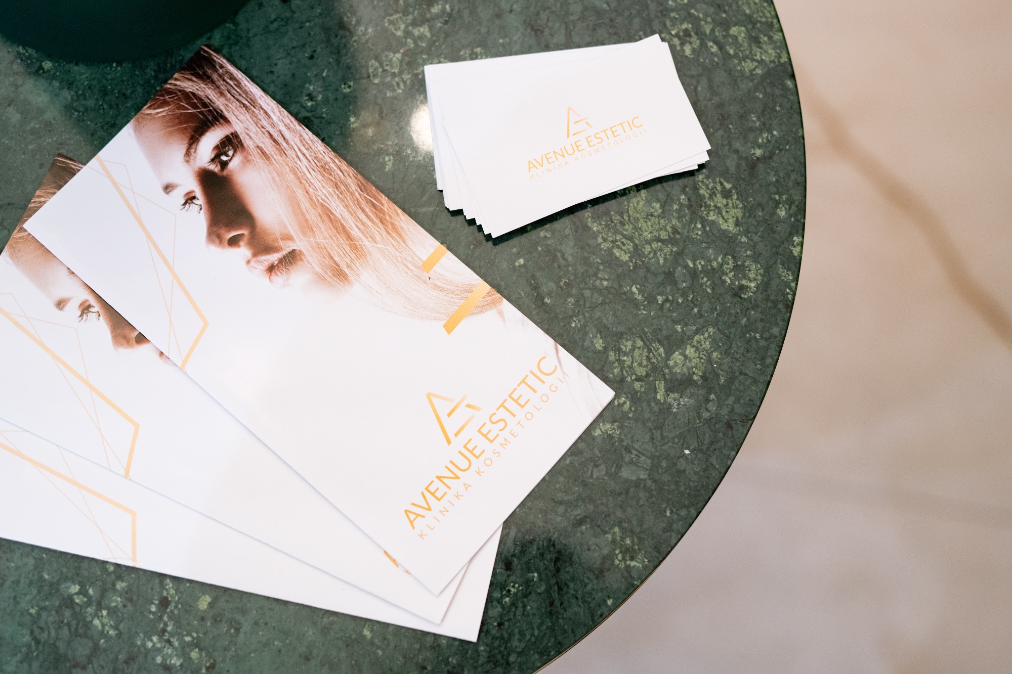 Ulotki i wizytówki z logo 'Avenue Estetic Klinika Kosmetologii' leżące na zielonym marmurowym stoliku.