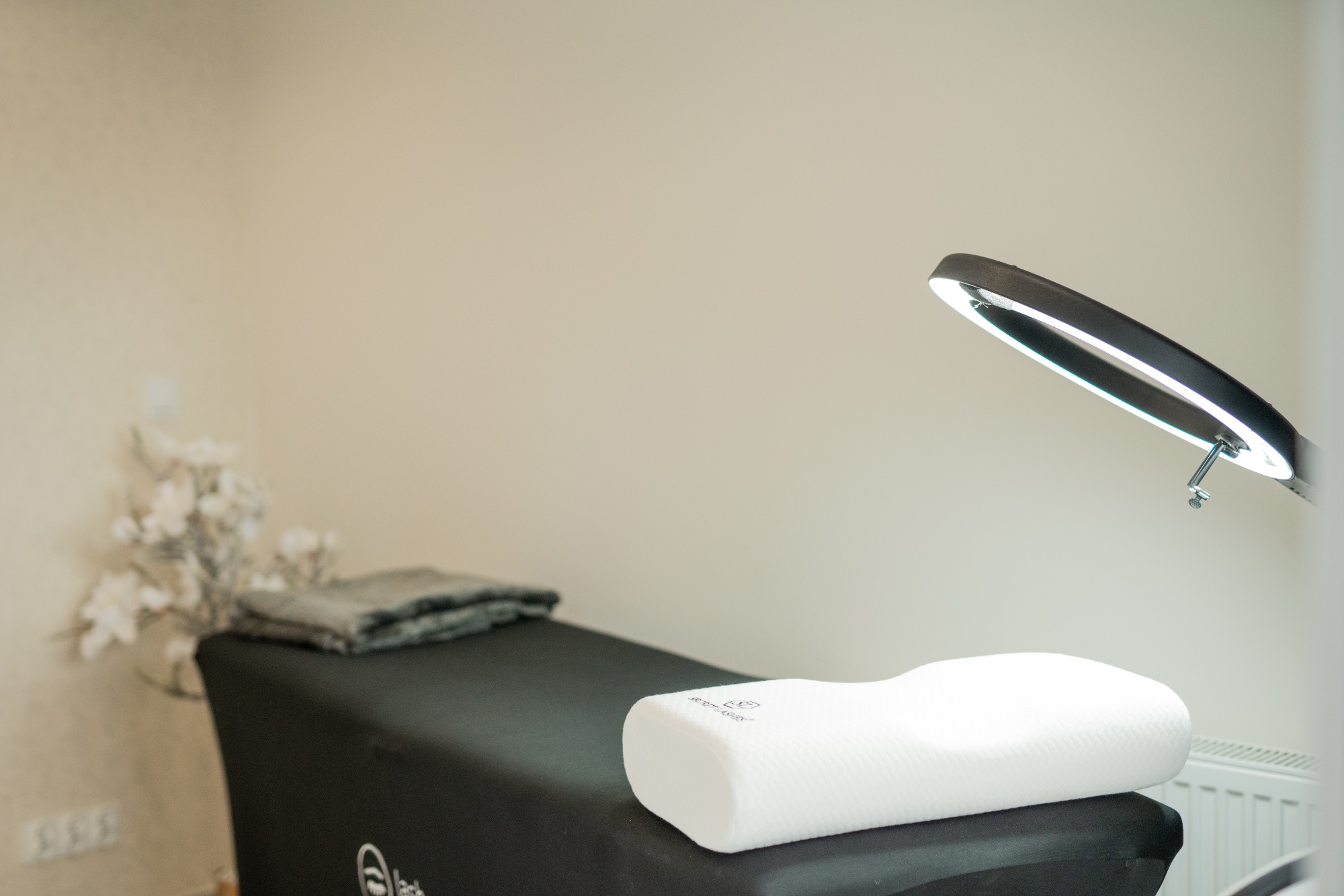 Przygotowane stanowisko zabiegowe z czarnym leżanką, białą poduszką ortopedyczną, złożonym szarym ręcznikiem i lampą pierścieniową w tle jasnych ścian i dekoracyjnych kwiatów.