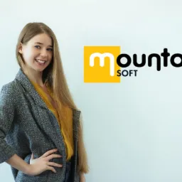 Uśmiechnięta młoda kobieta w szarej marynarce i żółtej bluzce, pozująca na tle białej ściany z logo firmy Mountain Soft w prawym górnym rogu.