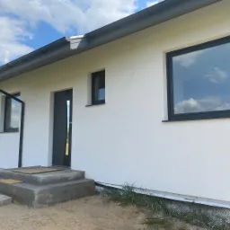 Wykończony dom jednorodzinny z białą elewacją, grafitowymi oknami i drzwiami, widoczne schody wejściowe i fragment dachu z rynnami, błękitne niebo z chmurami odbija się w szybach.