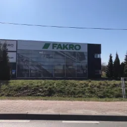 Budynek firmy Fakro z dużymi oknami wystawowymi prezentującymi okna dachowe i pionowe, widok z zewnątrz z chodnika i zielenią w tle.