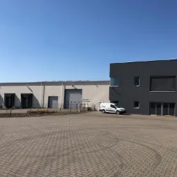 Hala magazynowa z rampami załadunkowymi i przylegający budynek biurowy z reklamą schodów strychowych, biała furgonetka parkuje na brukowanym placu przed budynkiem.