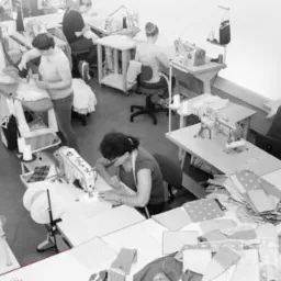 Widok z g&oacute;ry na szwalnię: kilka os&oacute;b pracuje przy maszynach do szycia, na stołach leżą stosy materiał&oacute;w, w tle widać manekina z suknią.