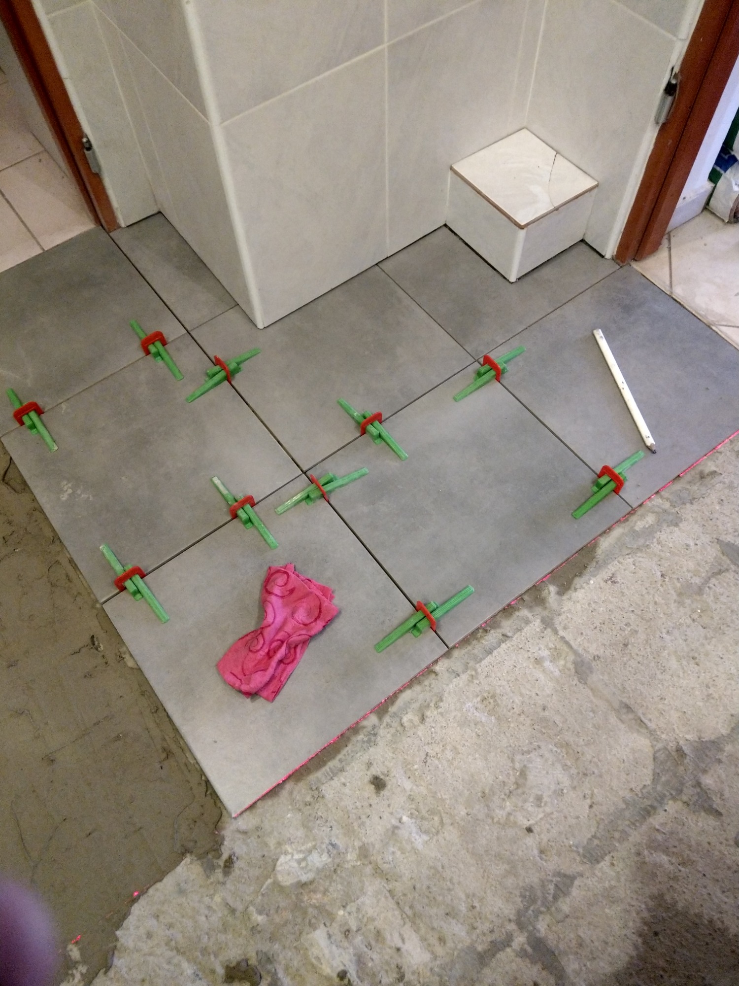 Układanie szarych płytek podłogowych w łazience, z użyciem zielonych klipsów poziomujących i czerwonych klinów, obok leży różowa ściereczka.