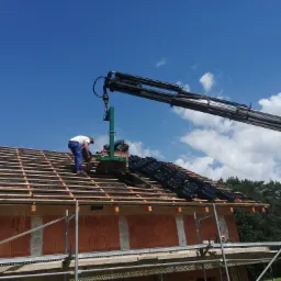 Układanie ciemnej dachówki na nowym dachu domu jednorodzinnego przy użyciu specjalistycznego podnośnika z chwytakiem, widok z dołu na rusztowanie i fragment lasu w tle.