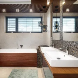 Nowoczesna łazienka z drewnianymi elementami: obudowa wanny z ciemnego drewna, blat pod umywalkami z jasnego drewna, mozaika na ścianie, wiszące lampy o geometrycznym kształcie, okno z żaluzjami.