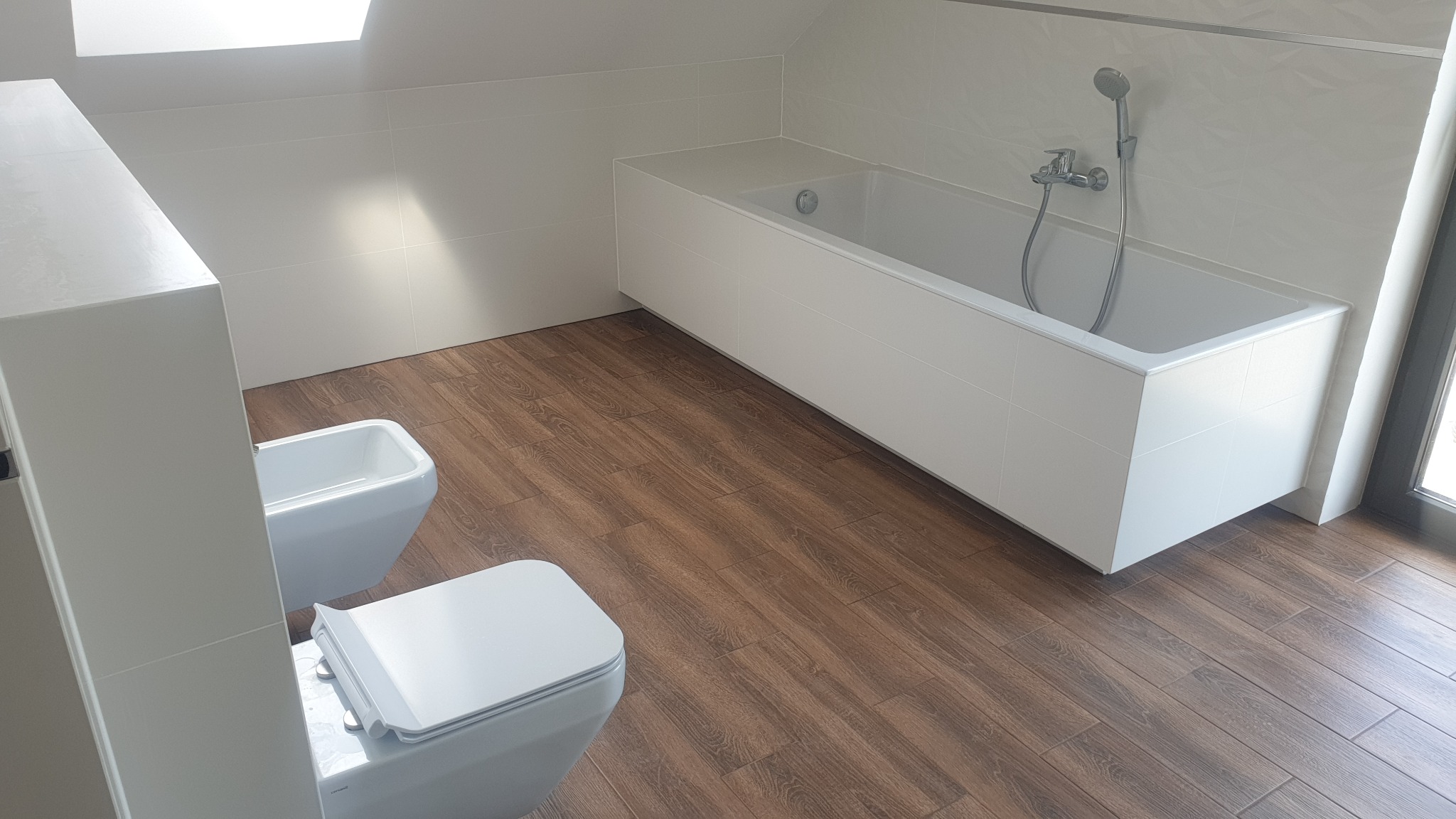 Nowoczesna łazienka na poddaszu z drewnopodobną podłogą, białą wanną obudowaną płytkami, toaletą i bidetem w minimalistycznym stylu, oświetlona światłem dziennym z okna dachowego.