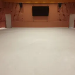 Jasna, gładka podłoga w przestronnym pomieszczeniu z brązowymi ścianami z pionowymi wgłębieniami, czarnym ekranem na ścianie i dwoma drzwiami po bokach.