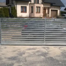 Nowoczesna, metalowa brama przesuwna z poziomymi szczeblami, widok z zewnątrz posesji, na tle domu jednorodzinnego.