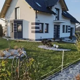 Nowoczesny dom jednorodzinny z białą elewacją, ciemną dachówką i szarymi akcentami dekoracyjnymi, otoczony zadbanym trawnikiem i ozdobnymi nasadzeniami.