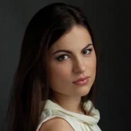 Portret młodej kobiety o ciemnych włosach i brązowych oczach, ubrana w jasną bluzkę na tle ciemnoszarego tła.