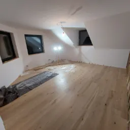 Jasne panele podłogowe ułożone w pokoju na poddaszu, widoczne okna dachowe, lampa budowlana i materiały zabezpieczające podłogę.