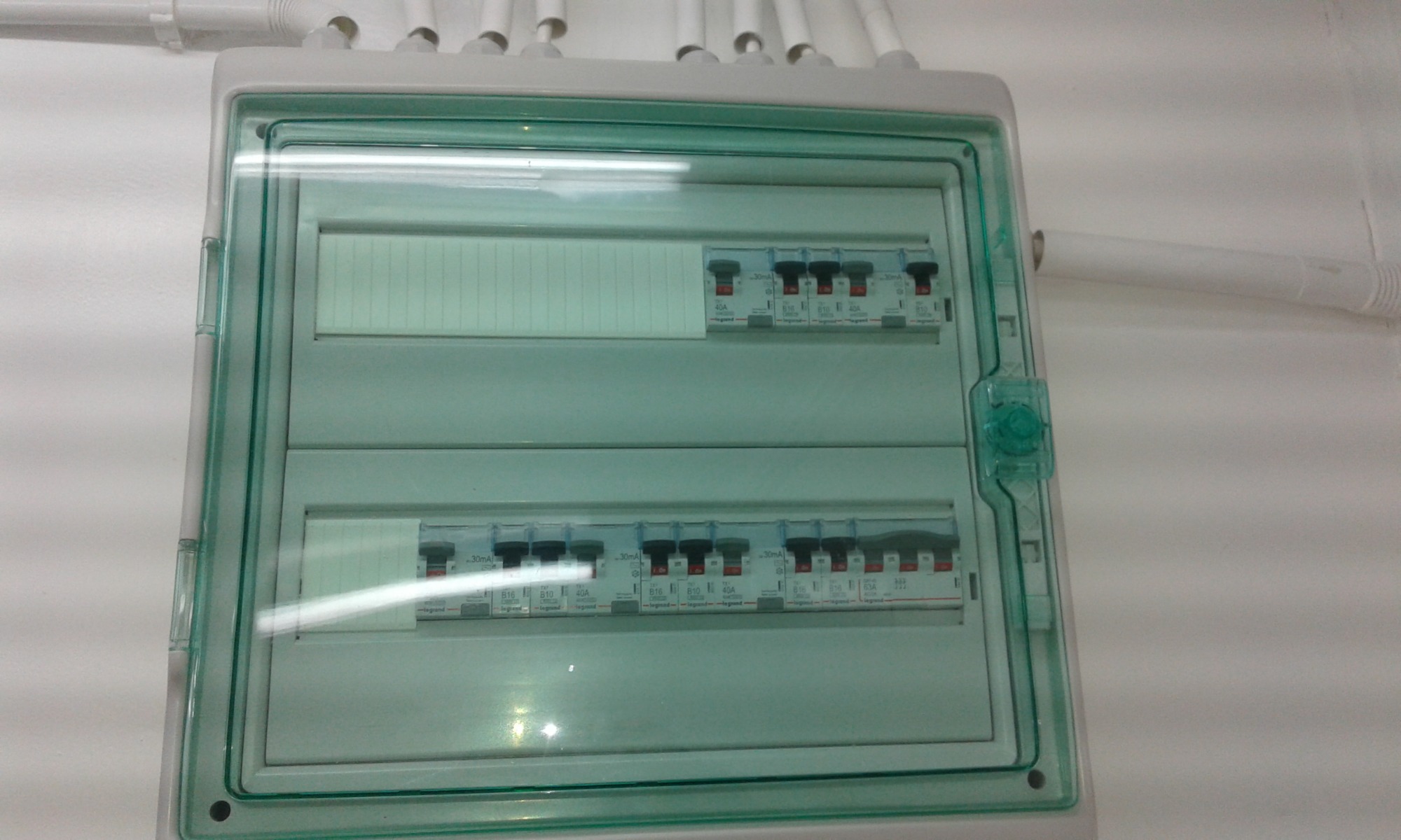 Rozdzielnica elektryczna z bezpiecznikami i przezroczystą, zielonkawą obudową, zamontowana na białej ścianie, widoczne rurki instalacyjne pod sufitem.