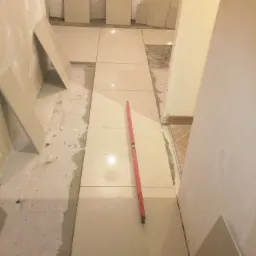 Układanie beżowych płytek podłogowych w korytarzu, widoczna poziomica i zapasowe płytki, surowy stan ścian.