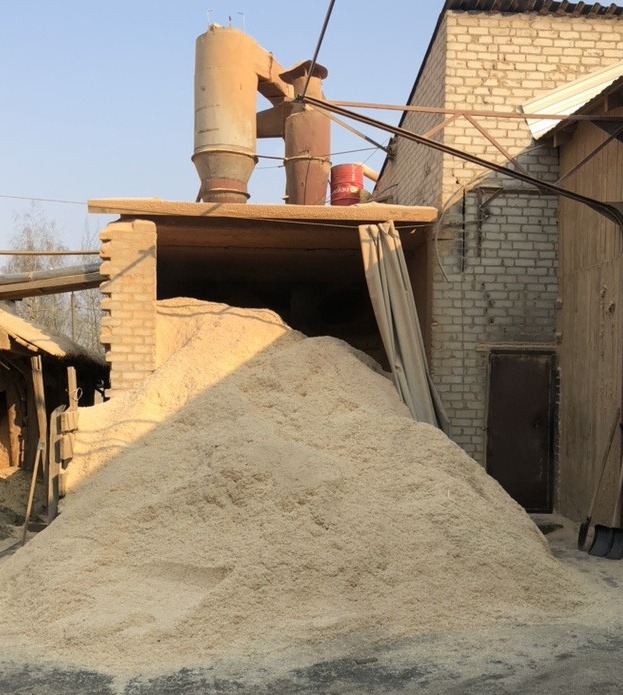 Góra jasnych wiórów pod zadaszeniem przy ceglanym budynku z instalacją przemysłową i czerwoną beczką na dachu.