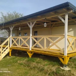 Zadaszony taras z drewnianą balustradą w kształcie krzyży, żółtą podłogą i schodami, oświetlony wiszącymi lampionami, widok z zewnątrz.