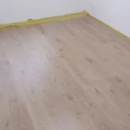 Jasna podłoga laminowana ułożona w pokoju, zabezpieczona żółtą folią malarską przy ścianach.