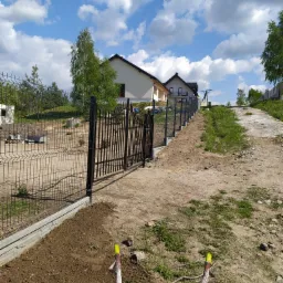 Montaż ogrodzenia panelowego z bramą wjazdową na nierównym terenie, widoczne słupki z żółtymi zaślepkami, w tle dom jednorodzinny pod błękitnym niebem.