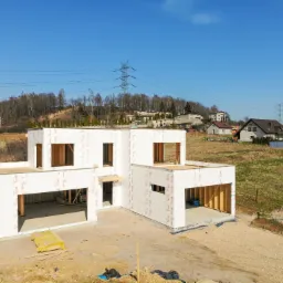 Surowy stan budynku modułowego z widoczną konstrukcją szkieletową, zabezpieczoną membraną Tyvek, na tle pagórkowatego terenu z liniami wysokiego napięcia.