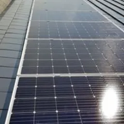 Rząd paneli słonecznych na dachu pokrytym ciemną dachówką, widoczne odbicie słońca na powierzchni paneli.