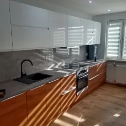 Nowoczesna kuchnia z białymi szafkami górnymi i drewnopodobnymi dolnymi, czarny zlew i bateria, szary panel nad blatem, drewniana podłoga, oświetlenie punktowe i rolety w oknie, z widocznymi...