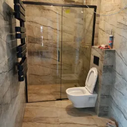 Wykończona łazienka z kabiną prysznicową typu walk-in, wiszącą toaletą i grzejnikiem drabinkowym na ścianie w beżowych płytkach imitujących marmur. Widoczny silikon i narzędzia.