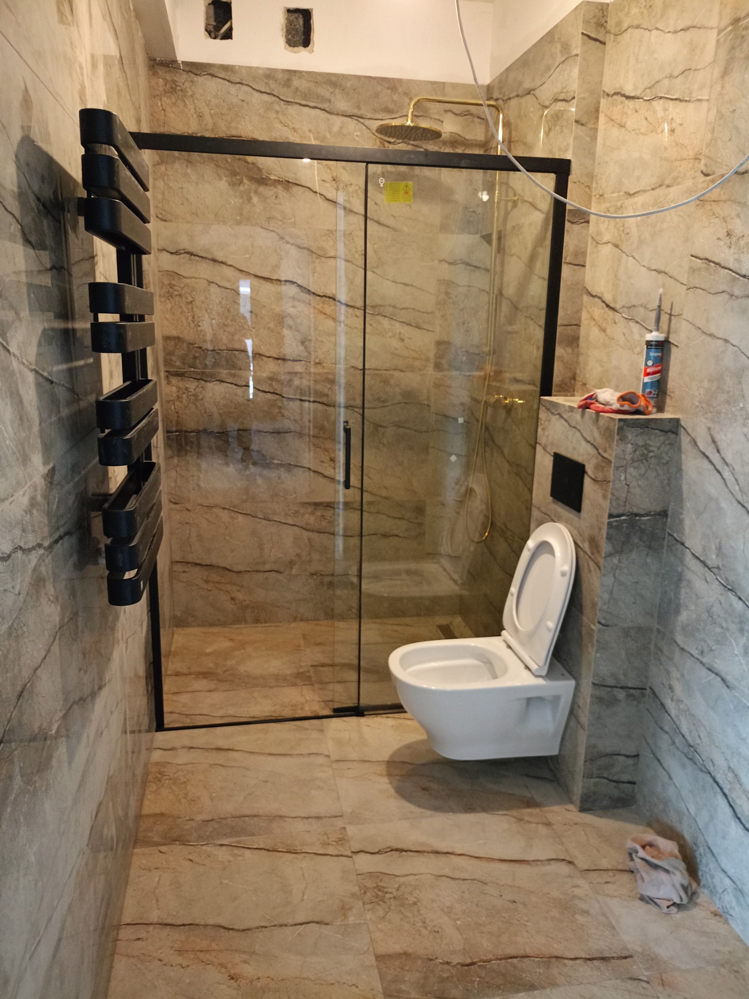 Wykończona łazienka z kabiną prysznicową typu walk-in, wiszącą toaletą i grzejnikiem drabinkowym na ścianie w beżowych płytkach imitujących marmur. Widoczny silikon i narzędzia.