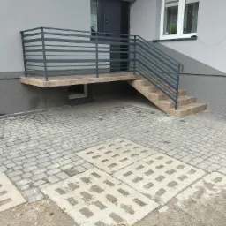 Metalowe schody zewnętrzne z poziomą balustradą prowadzące do drzwi wejściowych, wykończone płytkami, z widoczną kostką brukową na podjeździe.