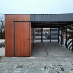 Nowoczesna wiata garażowa z metalową konstrukcją i panelowym ogrodzeniem w kolorze drewna, zamykana na furtkę, na tle szarej kostki brukowej i zimowego krajobrazu.