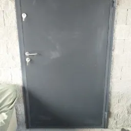 Szare, metalowe drzwi wejściowe z zamkiem i klamką, zamontowane w ścianie z białego kamienia dekoracyjnego.