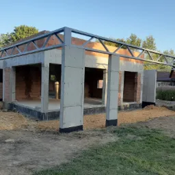 Konstrukcja z betonu komórkowego i metalu w trakcie budowy domu jednorodzinnego, widoczne cegły i piasek w otoczeniu.