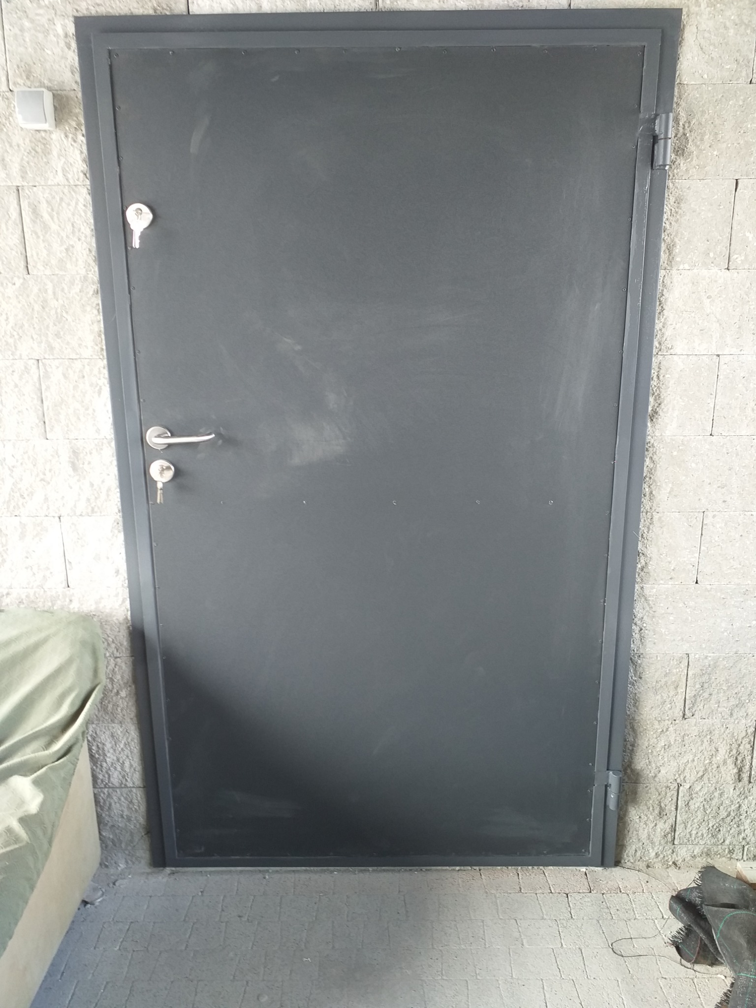 Szare, metalowe drzwi wejściowe z zamkiem i klamką, zamontowane w ścianie z białego kamienia dekoracyjnego.