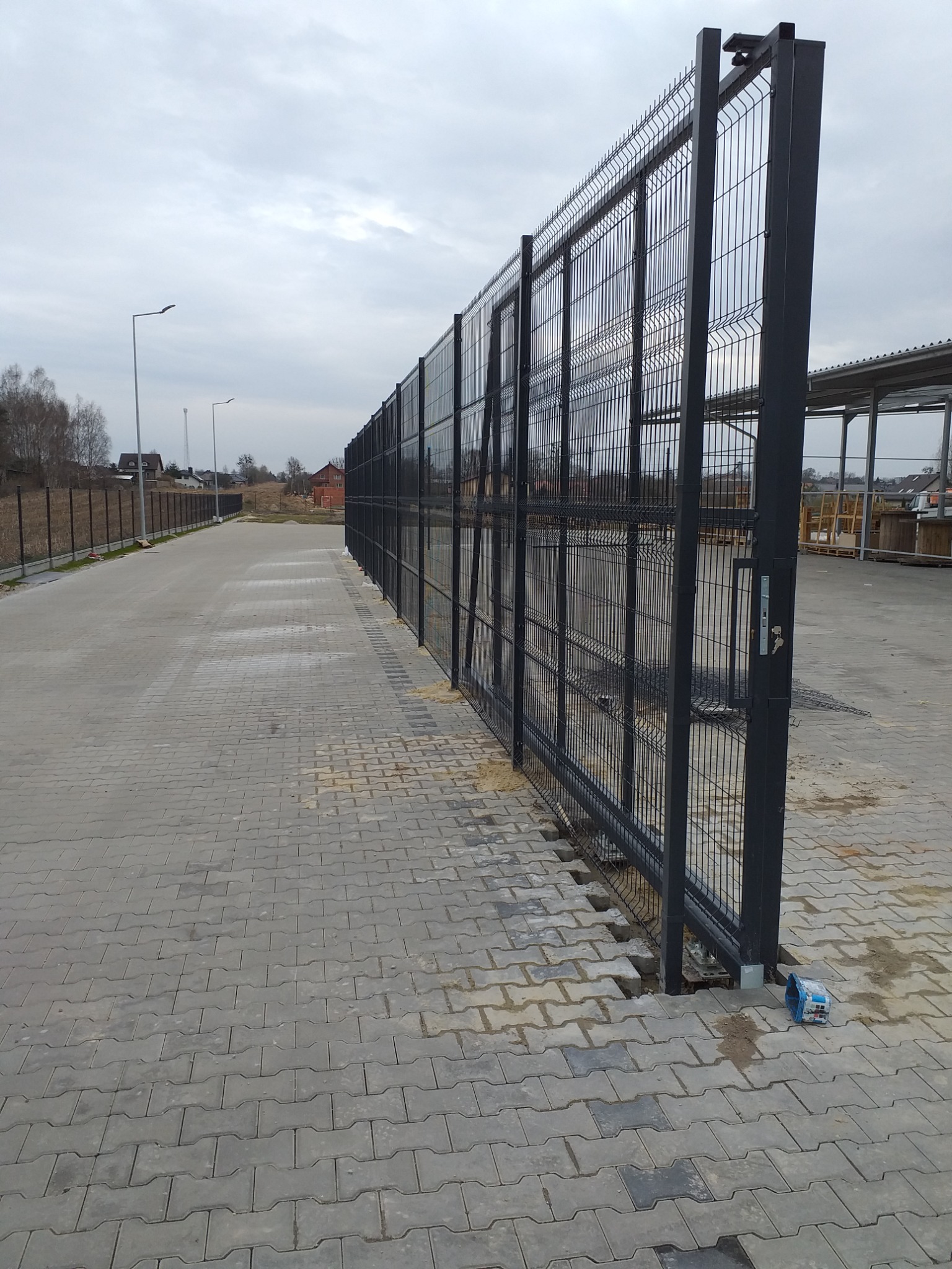 Długi, szary system ogrodzeniowy z paneli metalowych, widoczny z boku na tle szarej kostki brukowej i pochmurnego nieba. Widoczne elementy mocujące i zamek w furtce.