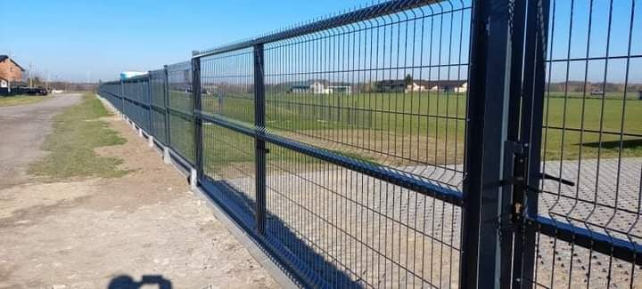 Długie, czarne ogrodzenie panelowe z furtką, zabezpieczające teren zielony, widoczne domy w oddali.