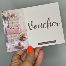 Strefa Piękna Beauty Salon - Dłoń trzymająca voucher do salonu kosmetycznego Strefa Piękna z motywem prezentów i napisem Voucher, na palcu pomarańczowy lakier hybrydowy z efektem lustra.