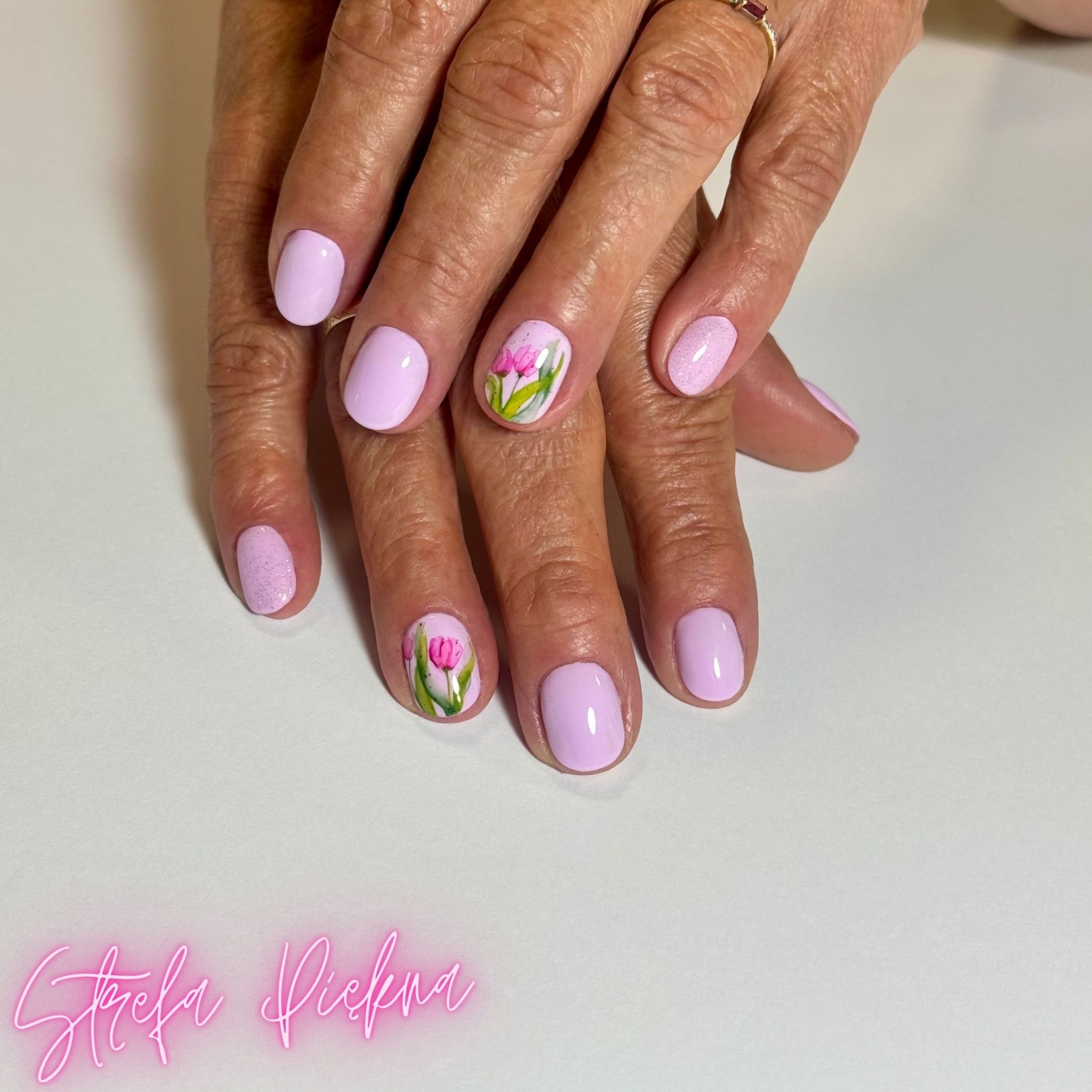 Dłonie z pastelowymi, liliowymi paznokciami, dwa z nich zdobione ręcznie malowanymi tulipanami, na białym tle z neonowym napisem 'Strefa Piękna' w rogu.